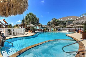 Una piscina con un deck circundante, palmeras y sombrillas de techo de paja.