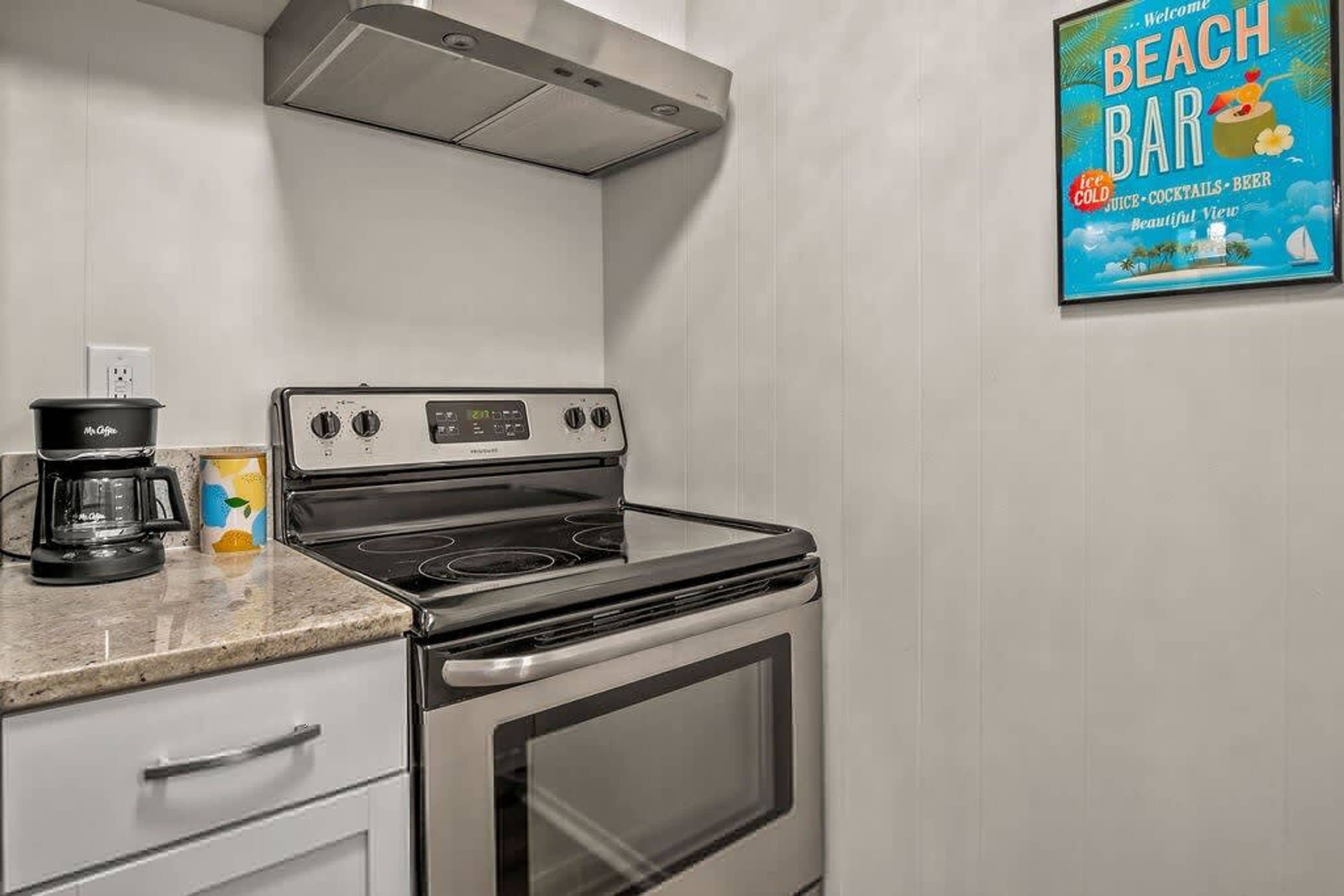 A kitchen with a stove, a coffee maker, and a wall-mounted sign advertising a beach bar.