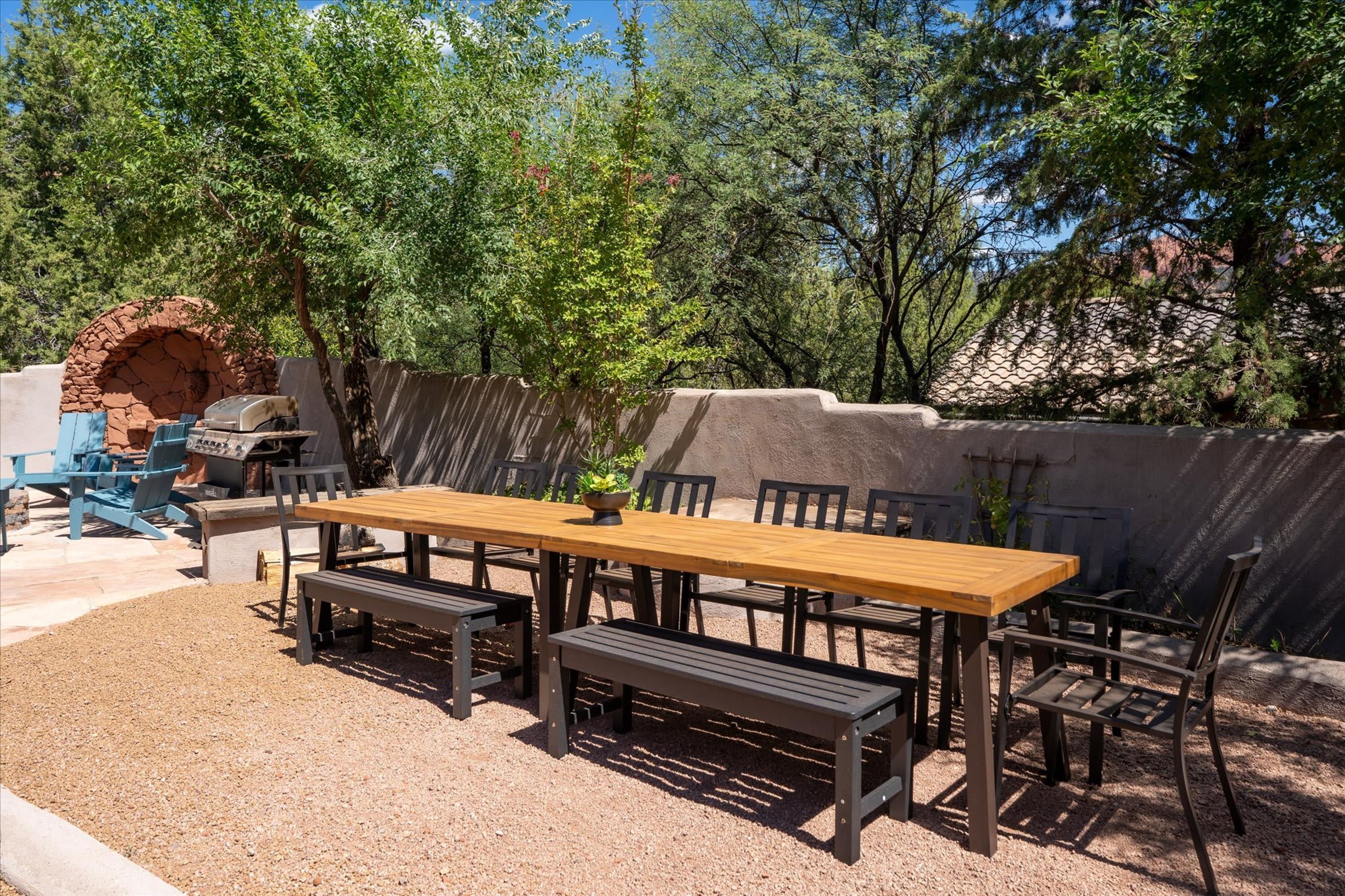 Un patio al aire libre con una mesa de madera y bancos, rodeado por un muro de piedra y árboles.