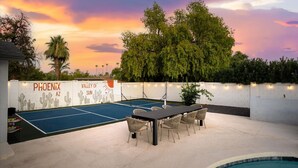 An outdoor tennis court with a pool table, surrounded by chairs and a dining table, with a sunset sky in the background. - Backyard Pickleball Hottub Pool Gameroom (Phoenix)