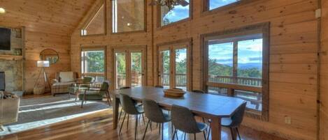 A spacious room with a large dining table and chairs, a fireplace, and large windows offering views of the outdoors.