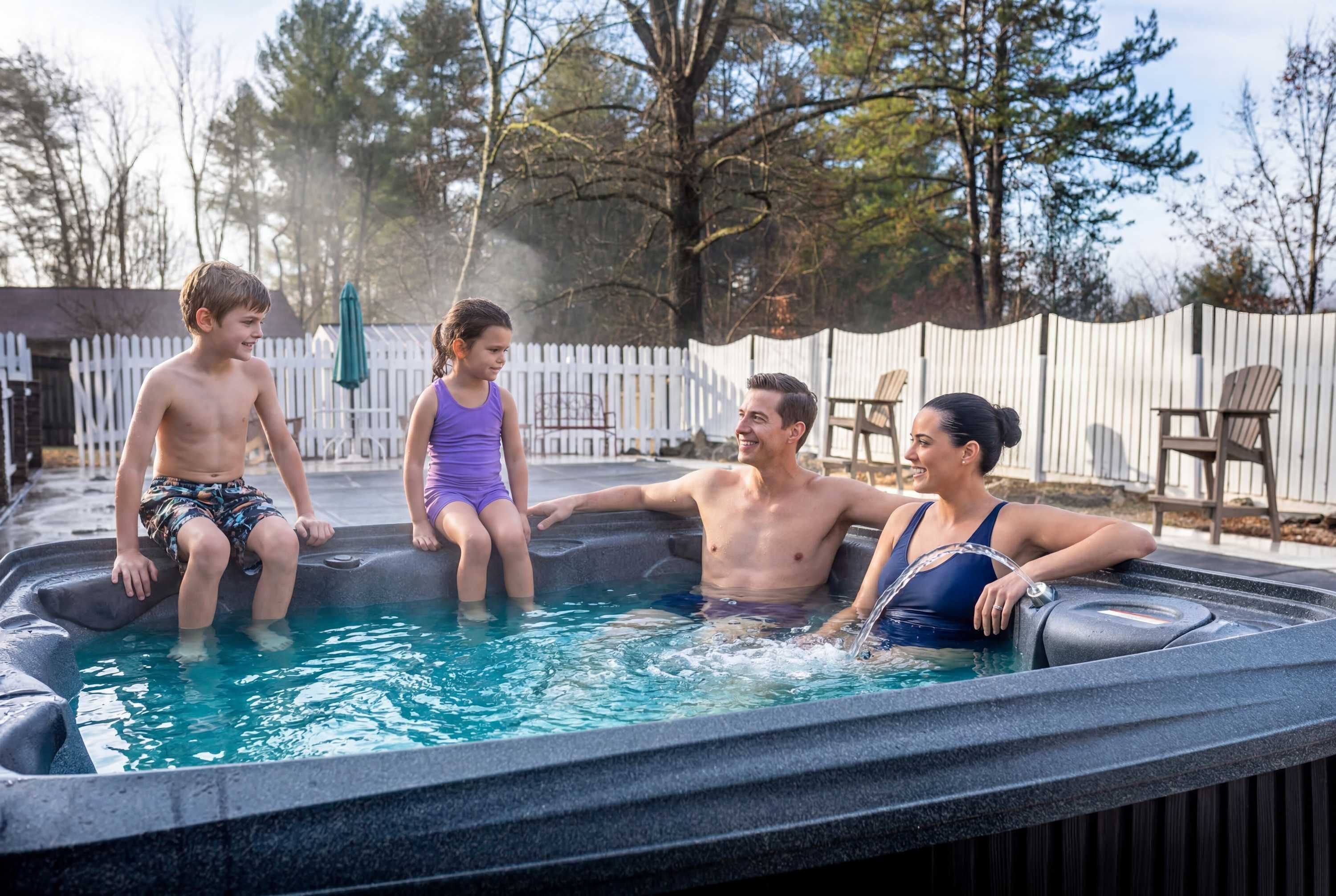 Une famille de quatre personnes dans un jacuzzi, avec une clôture blanche et des arbres en arrière-plan.