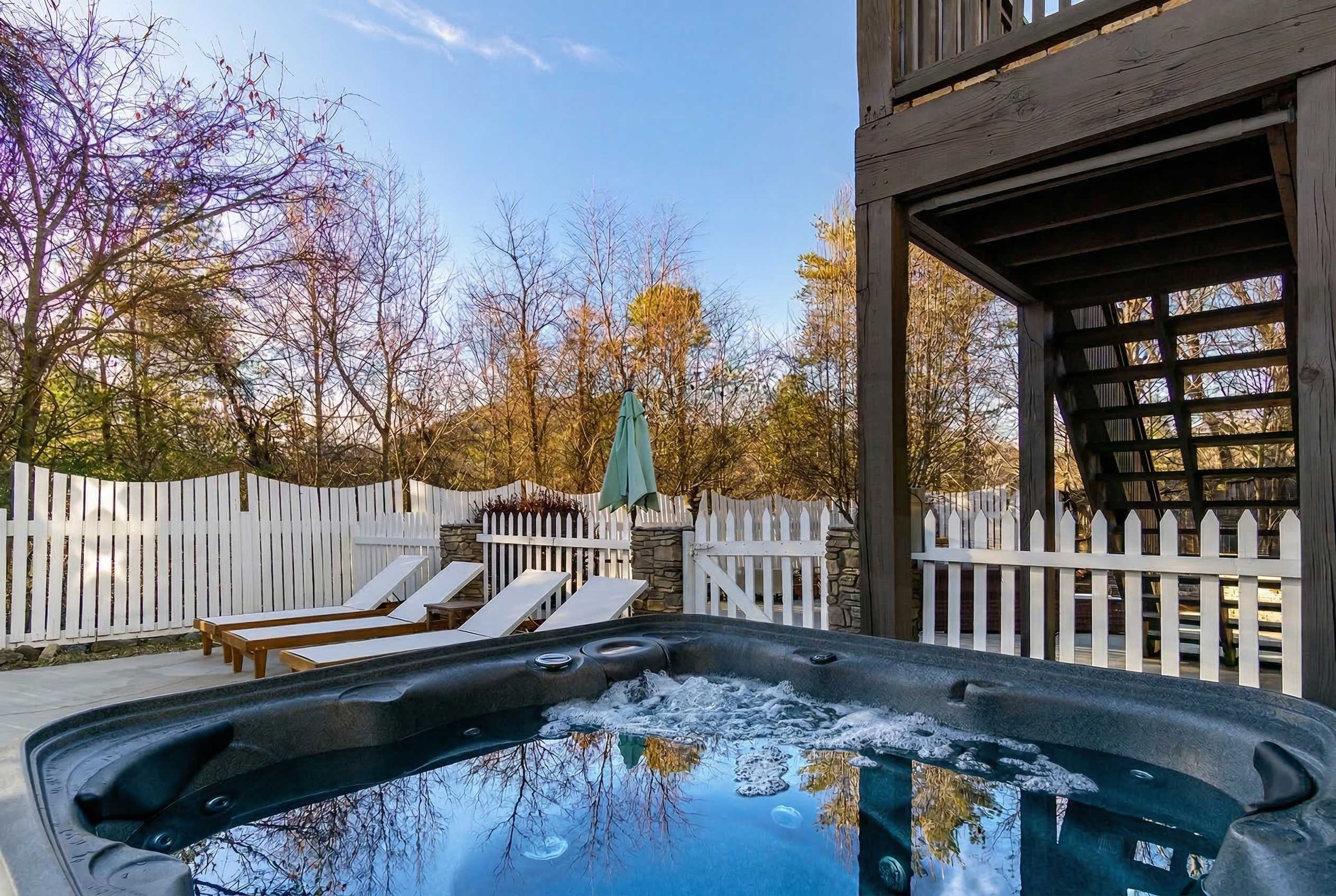 A hot tub with a wooden deck and lounge chairs, surrounded by a white picket fence and trees.
