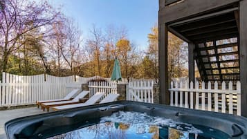 A hot tub with a wooden deck and lounge chairs, surrounded by a white picket fence and trees.