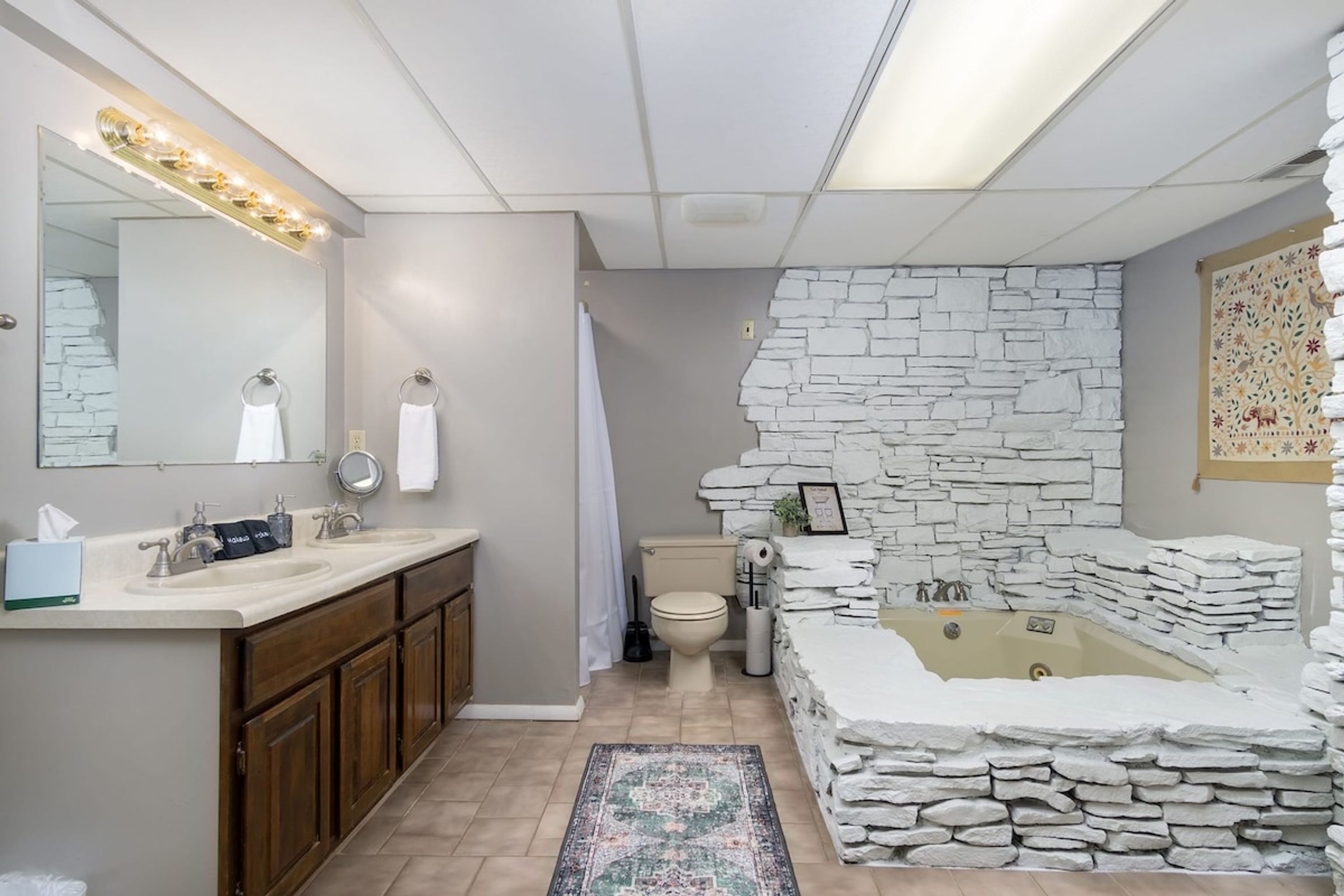 Een badkamer met een stenen muur, een badkuip, een toilet en een wastafel met spiegel en verlichting.