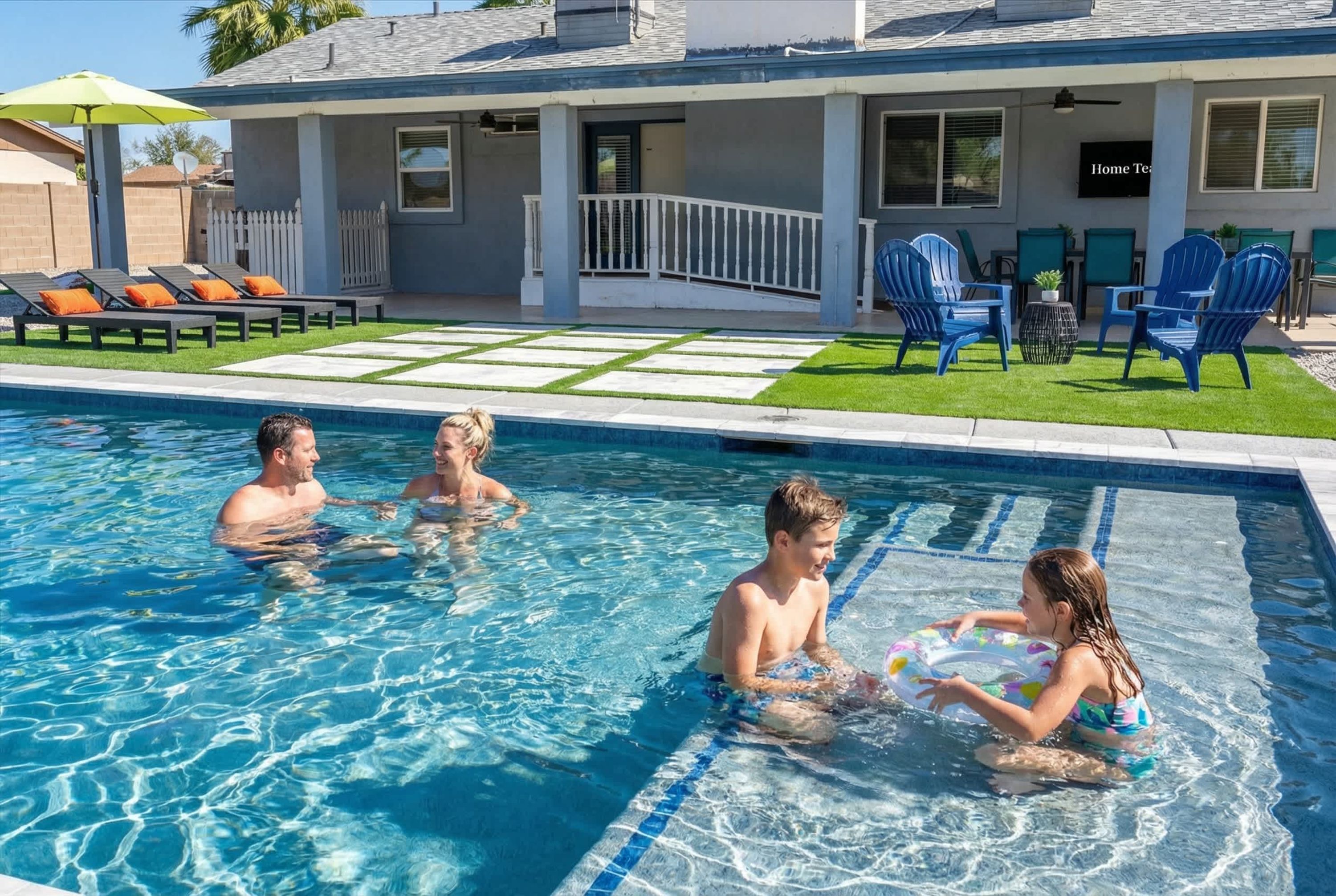 A swimming pool with a clear blue water surface, surrounded by a deck area with lounge chairs and a small patio with chairs and a table.