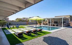 Un patio trasero con piscina, mobiliario exterior y una zona de barbacoa techada.