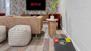 A living room with a flat-screen TV displaying 'NETFLIX,' a sofa, ottomans, a coffee table, a potted plant, and a small exercise mat with a towel and a water bottle. - The Palm Putt Heated Pool Golden Tee Bocce (Phoenix)