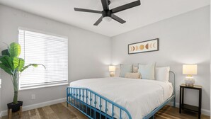 A bedroom with a bed, a ceiling fan, a window with blinds, a potted plant, and a wall-mounted lamp. - The Palm Putt Heated Pool Golden Tee Bocce (Phoenix)