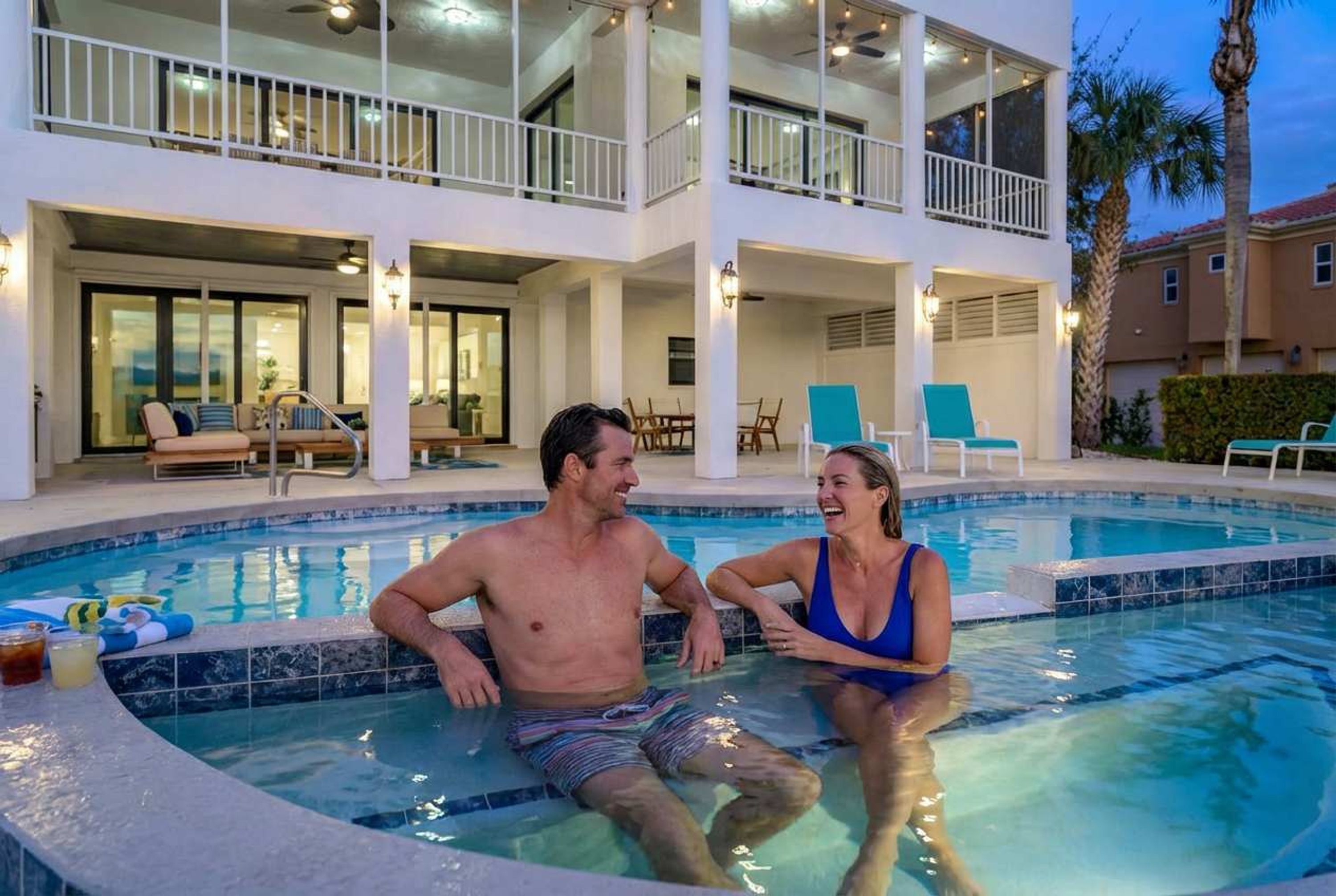A swimming pool area with a two-story building, lounge chairs, and a palm tree.