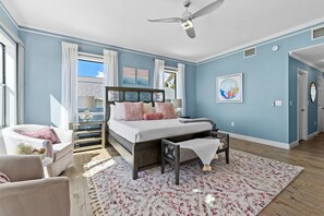 Ein Schlafzimmer mit einem Bett, einem Deckenventilator, einem Fenster mit Vorhängen, einem kleinen Tisch und einem Sessel.