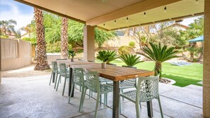 Ein überdachter Patio-Bereich mit Tisch und Stühlen, Palmen und Blick auf einen Garten mit Rasen und Pool.