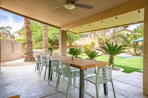 Ein überdachter Patio-Bereich mit Tisch und Stühlen, Palmen und Blick auf einen Garten mit Rasen und Pool.