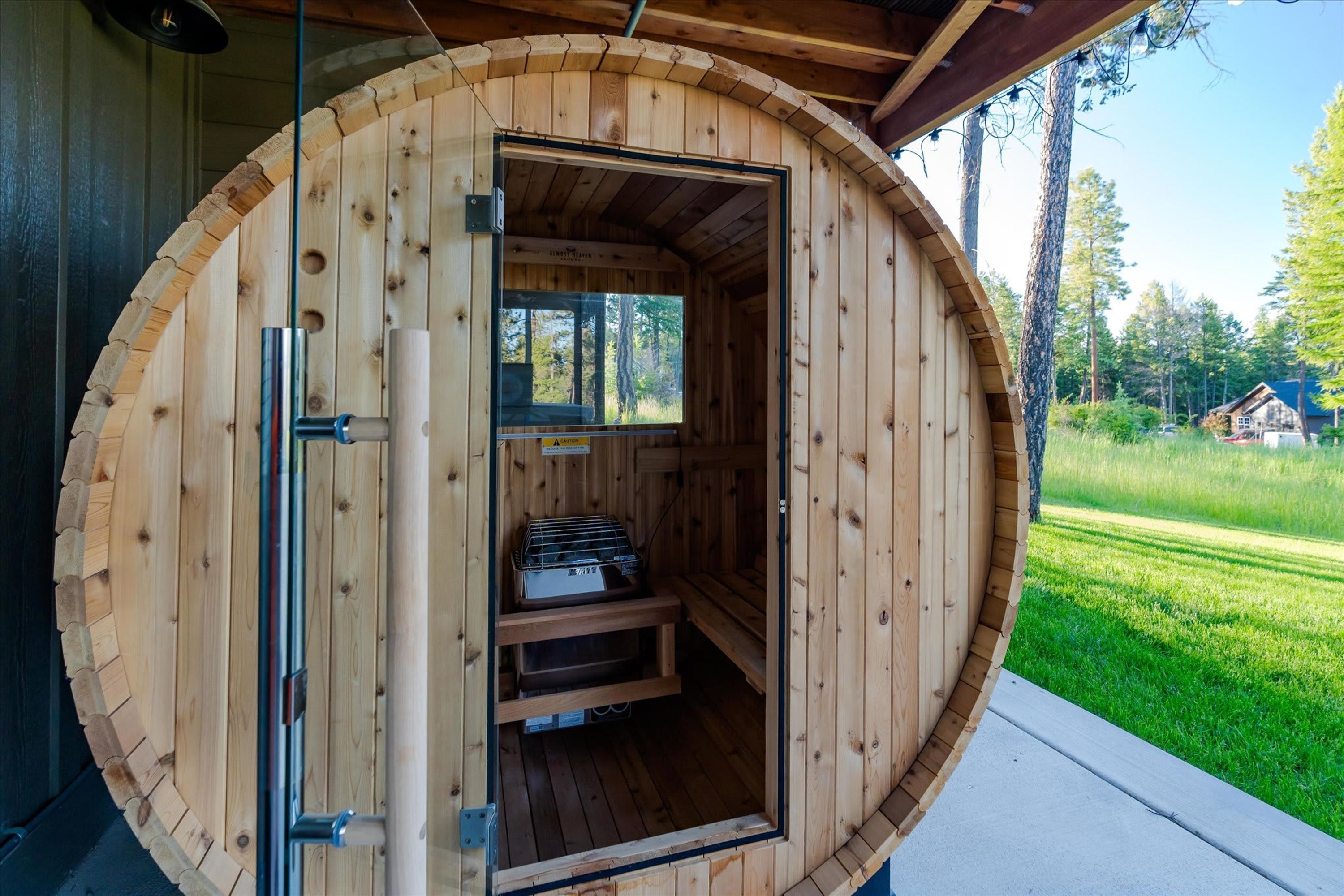 A wooden sauna with a glass door, a bench inside, and a view of the surrounding forest.
