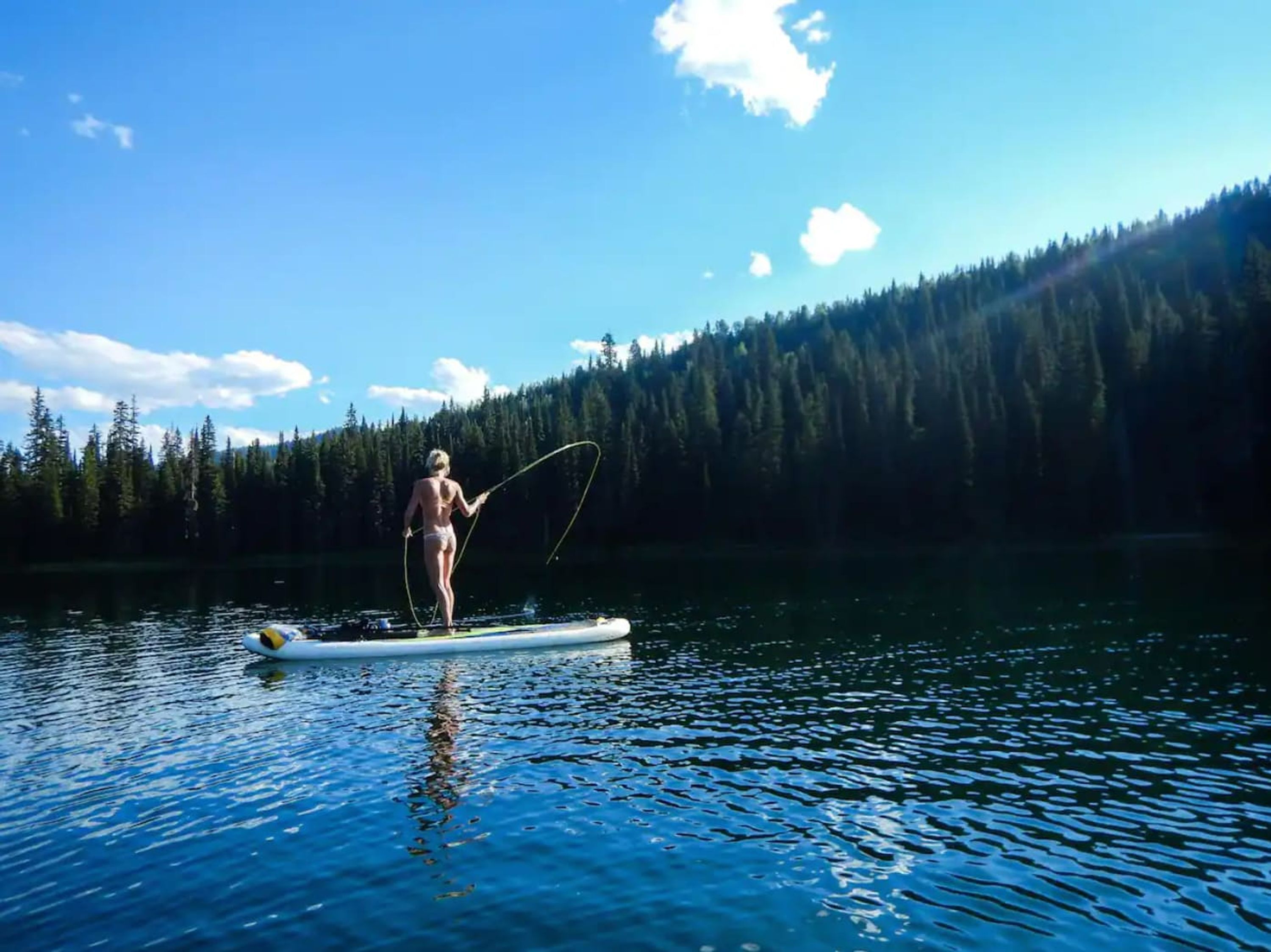 Eine Person steht auf einem Paddleboard auf einem ruhigen See, im Hintergrund ein bewaldeter Berg.