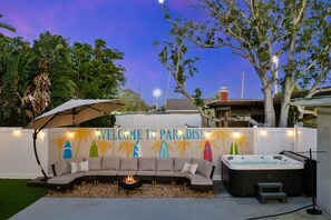 A patio area with a curved sofa, a fire pit, and a hot tub. A sign reads 'WELCOME TO PARADISE' with surfboard silhouettes. - 4 Br Oasis Pool Tiki Hut Hot Tub Ping Pong Arcade (Seminole)
