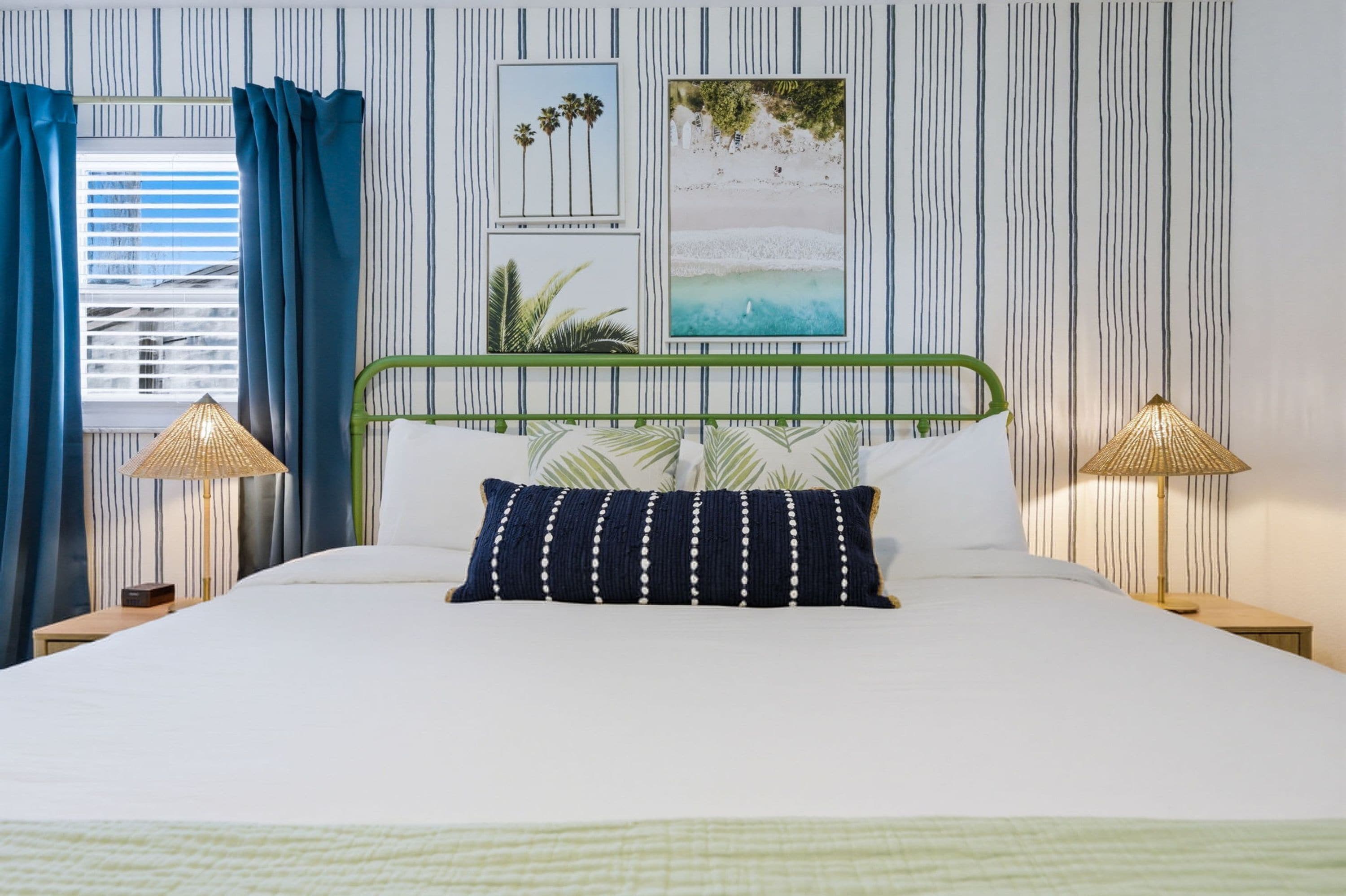 Un lit avec un cadre en métal vert, une literie blanche et un oreiller bleu foncé à pois blancs. Il y a deux lampes de chevet avec un abat-jour en osier. Le mur est décoré de rayures verticales et de deux tableaux encadrés.