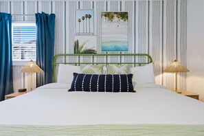 A bed with a green metal frame, white bedding, and a dark blue pillow with white dots. There are two bedside lamps with a woven shade. The wall has vertical stripes and two framed pictures. - 4 Br Oasis Pool Tiki Hut Hot Tub Ping Pong Arcade (Seminole)