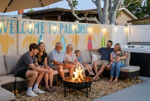 A group of people seated around a fire pit in an outdoor setting with a 'WELCOME TO PARADISE' sign in the background. - 4 Br Oasis Pool Tiki Hut Hot Tub Ping Pong Arcade (Seminole)
