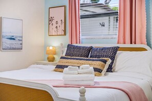 A neatly made bed with pillows and a folded towel, a bedside lamp, a framed picture on the wall, and a window with curtains. - 4 Br Oasis Pool Tiki Hut Hot Tub Ping Pong Arcade (Seminole)