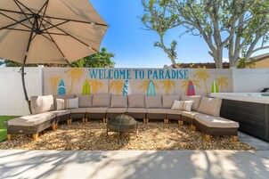 Un espacio exterior con asientos curvos y un letrero que dice 'WELCOME TO PARADISE', rodeado de palmeras y una cerca blanca.