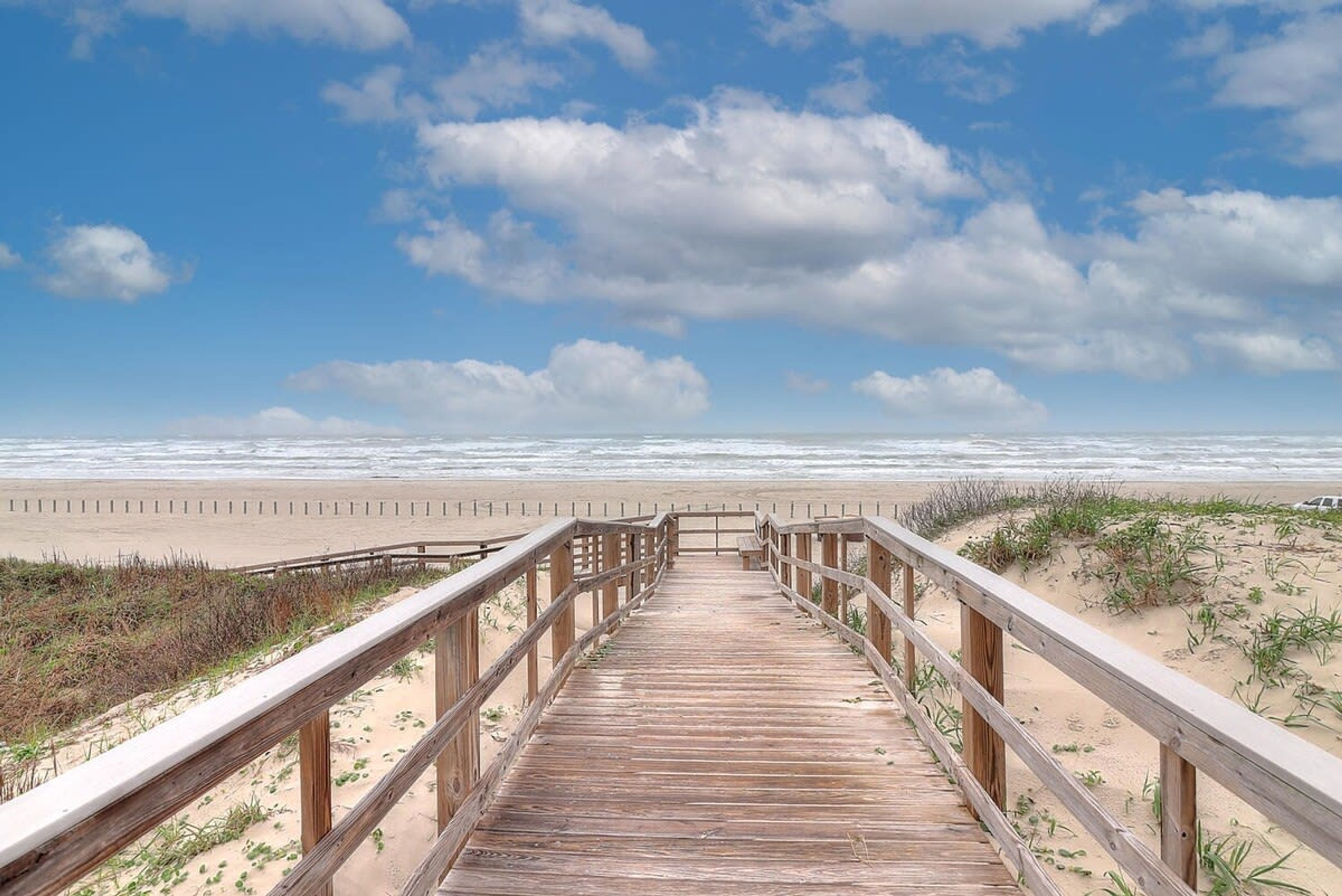 Une promenade en bois menant à une plage, avec un ciel dégagé et l’océan en arrière-plan.