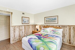 Ein Schlafzimmer mit hölzernem Kopfteil, einem Bett mit farbenfroher Blumenbettdecke, einem Nachttisch mit Lampe und zwei gerahmten Bildern an der Wand.