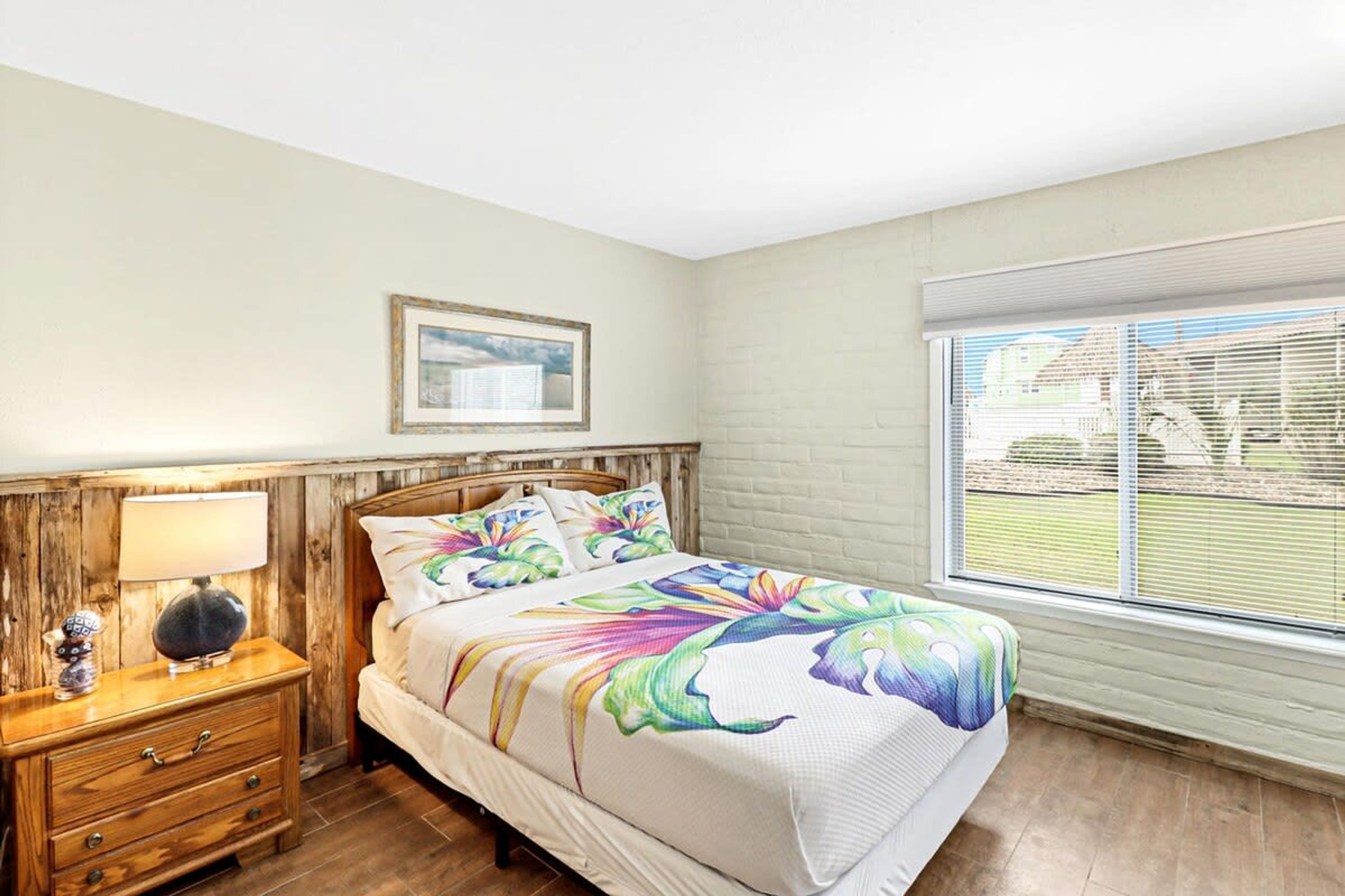 A bedroom with a bed, a nightstand, a lamp, a window with blinds, and a framed picture on the wall.