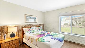 A bedroom with a bed, a nightstand, a lamp, a window with blinds, and a framed picture on the wall.