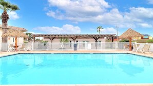A swimming pool with a pergola and lounge chairs, surrounded by a fence and palm trees. - Sea Escape-boardwalk Access Large Community Pool (Port Aransas)