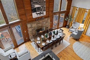 Un coin repas agrémenté d’une cheminée en pierre, d’une table à manger en bois et de chaises. Une grande fenêtre, ornée de rideaux, offre une vue sur l’extérieur.