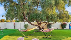 Ein gepflegter Rasen mit zwei Hängematten und zwei Poufs, im Hintergrund ein Golfplatz und ein großer Baum, der Schatten spendet.