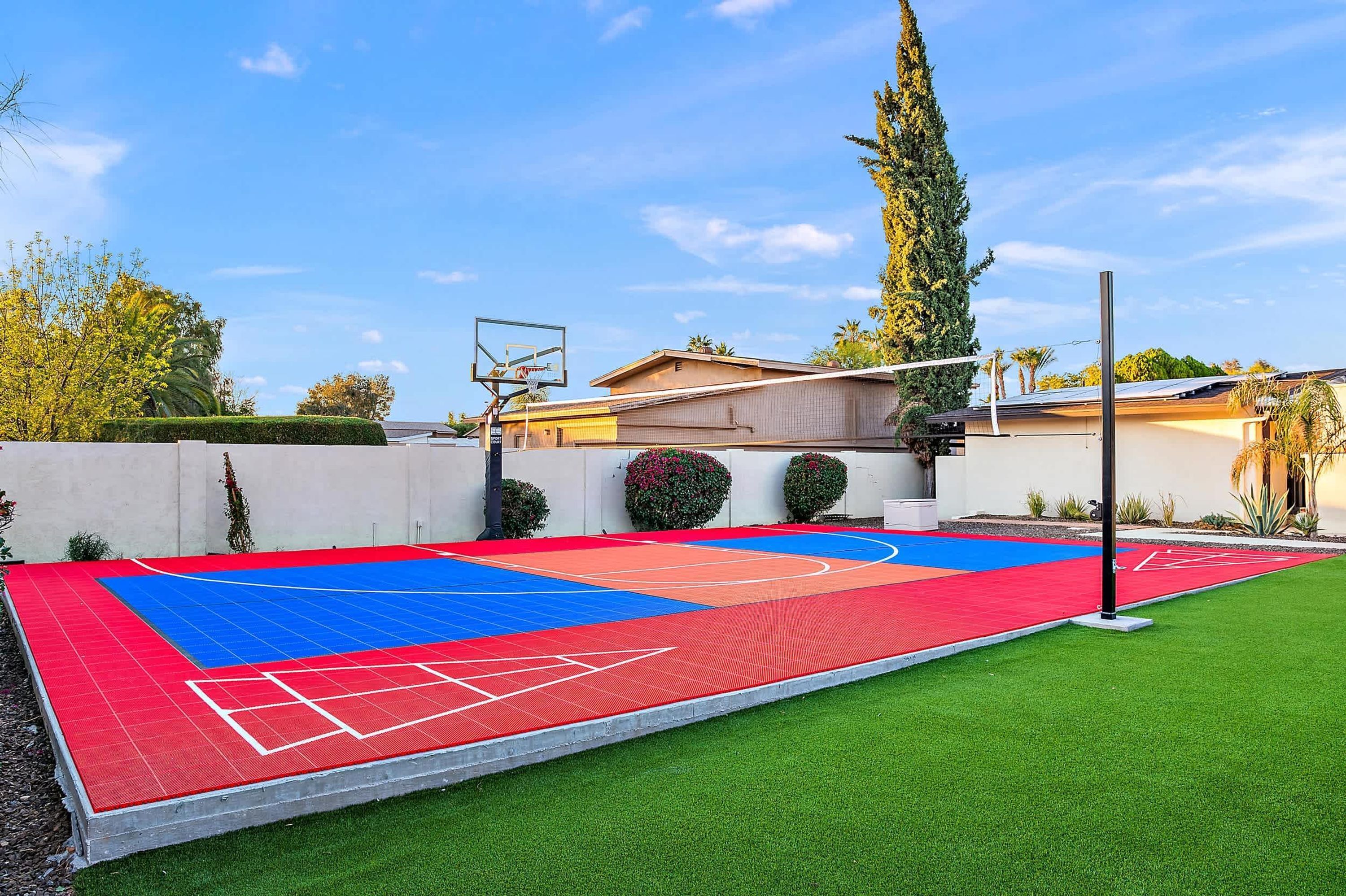 Una cancha de baloncesto al aire libre con superficie roja y azul, rodeada por un muro blanco y vegetación.