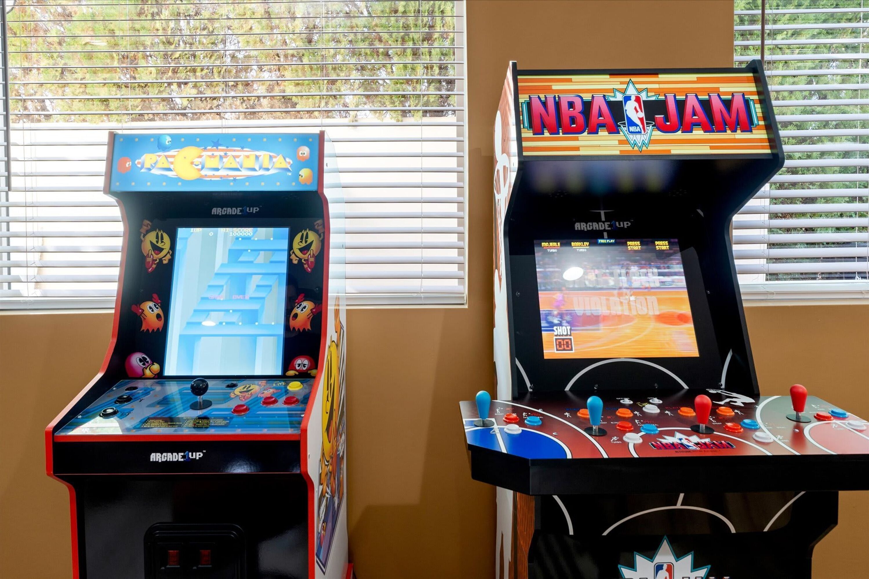 To arkademaskiner, hvoraf den ene er mærket 'Pacman' og den anden 'NBA Jam', begge med joystick og skærme der viser spilgrafik.