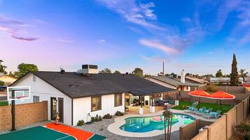 A residential backyard with a swimming pool, a basketball court, and a patio area with outdoor furniture.