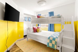 A bunk bed room with a television mounted on the wall, a window with curtains, and a yellow accent wall. - The Brightside Heated Pool Bball Court Bocce (Glendale)
