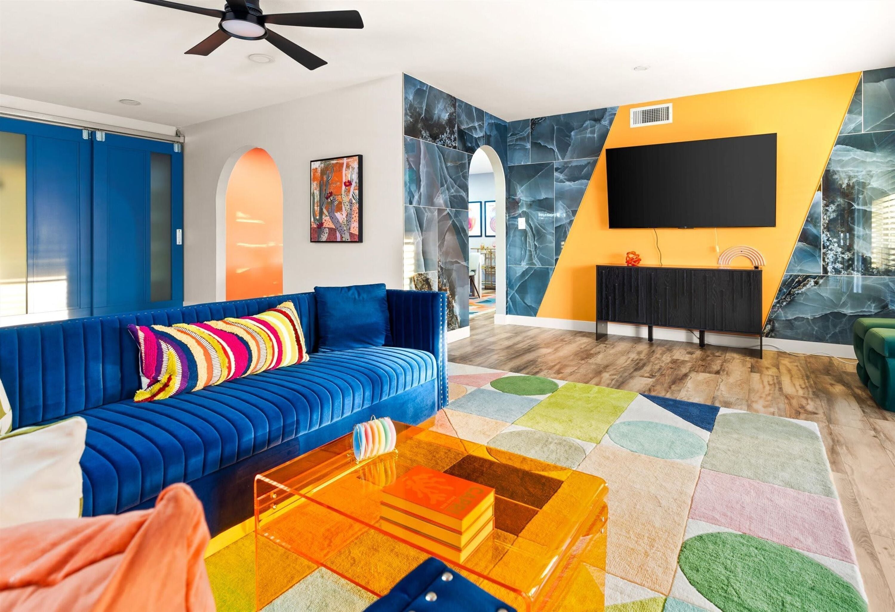 A modern living room with a blue sofa, a colorful rug, and a flat-screen TV mounted on a yellow wall.