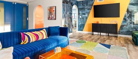 A modern living room with a blue sofa, a colorful rug, and a flat-screen TV mounted on a yellow wall.