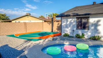 A backyard with a swimming pool, a basketball hoop, and a tennis court.