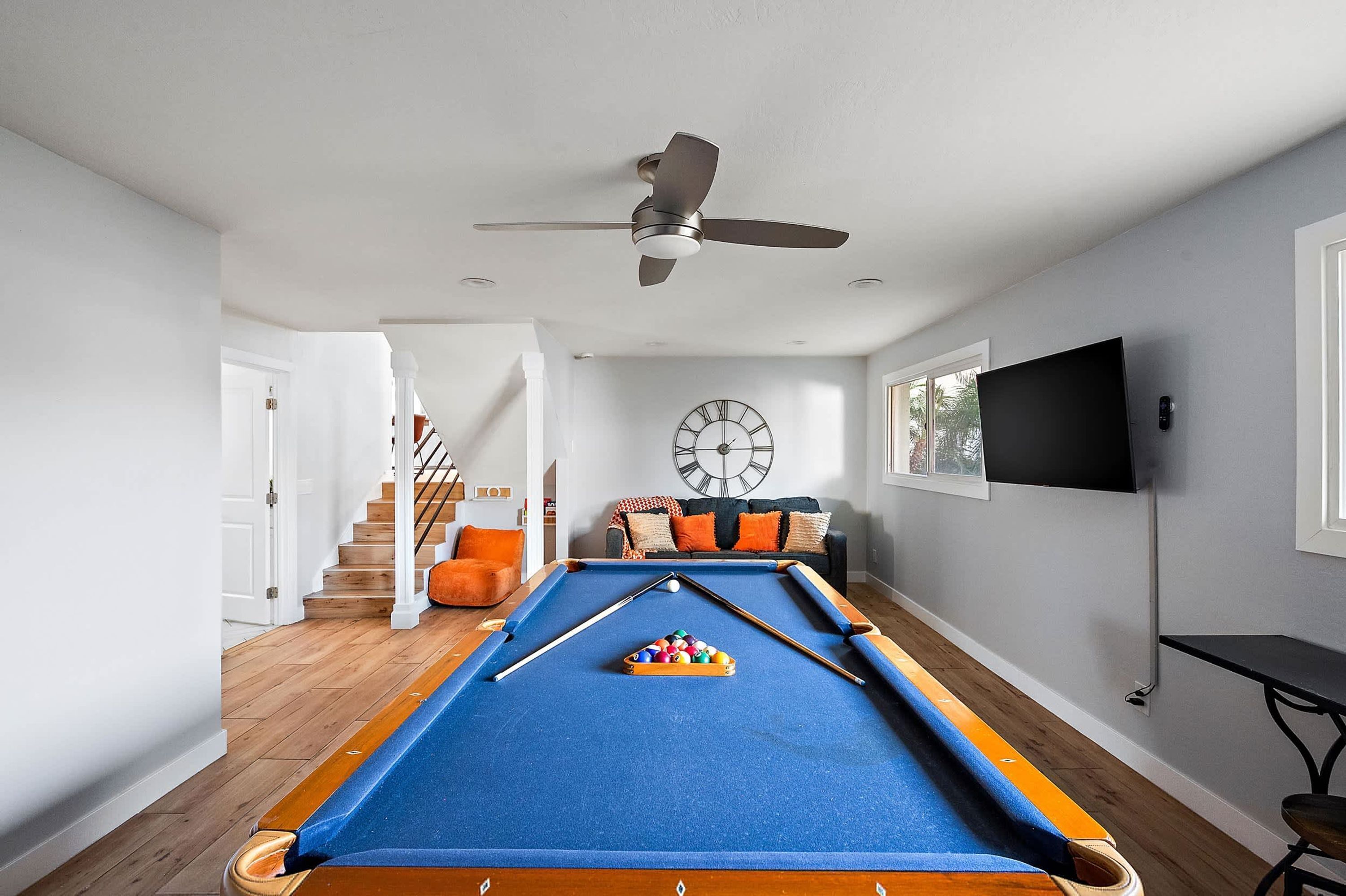 A pool table with a clock and a television in a room with a staircase and a window.