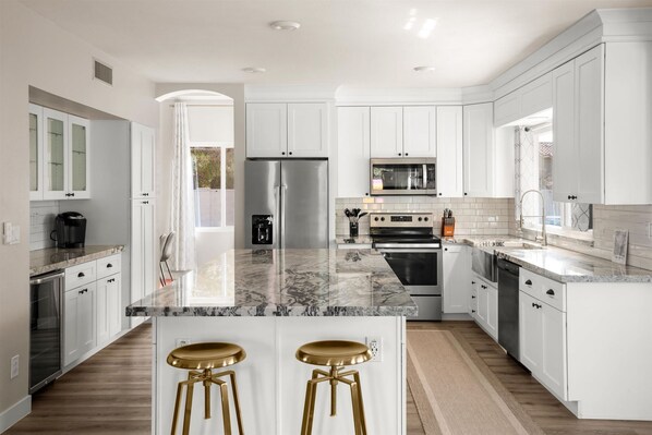 A modern kitchen with white cabinetry, a central island with stools, stainless steel appliances, and a view through an open door. - Private Pickleball Court Movie Room Hot Tub (Scottsdale)