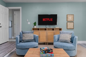 Ein Wohnzimmer mit zwei blauen Sesseln, einem Couchtisch mit einem Gesellschaftsspiel, einem Fernseher, der 'NETFLIX' anzeigt, und einem Schrank mit einer Pflanze.