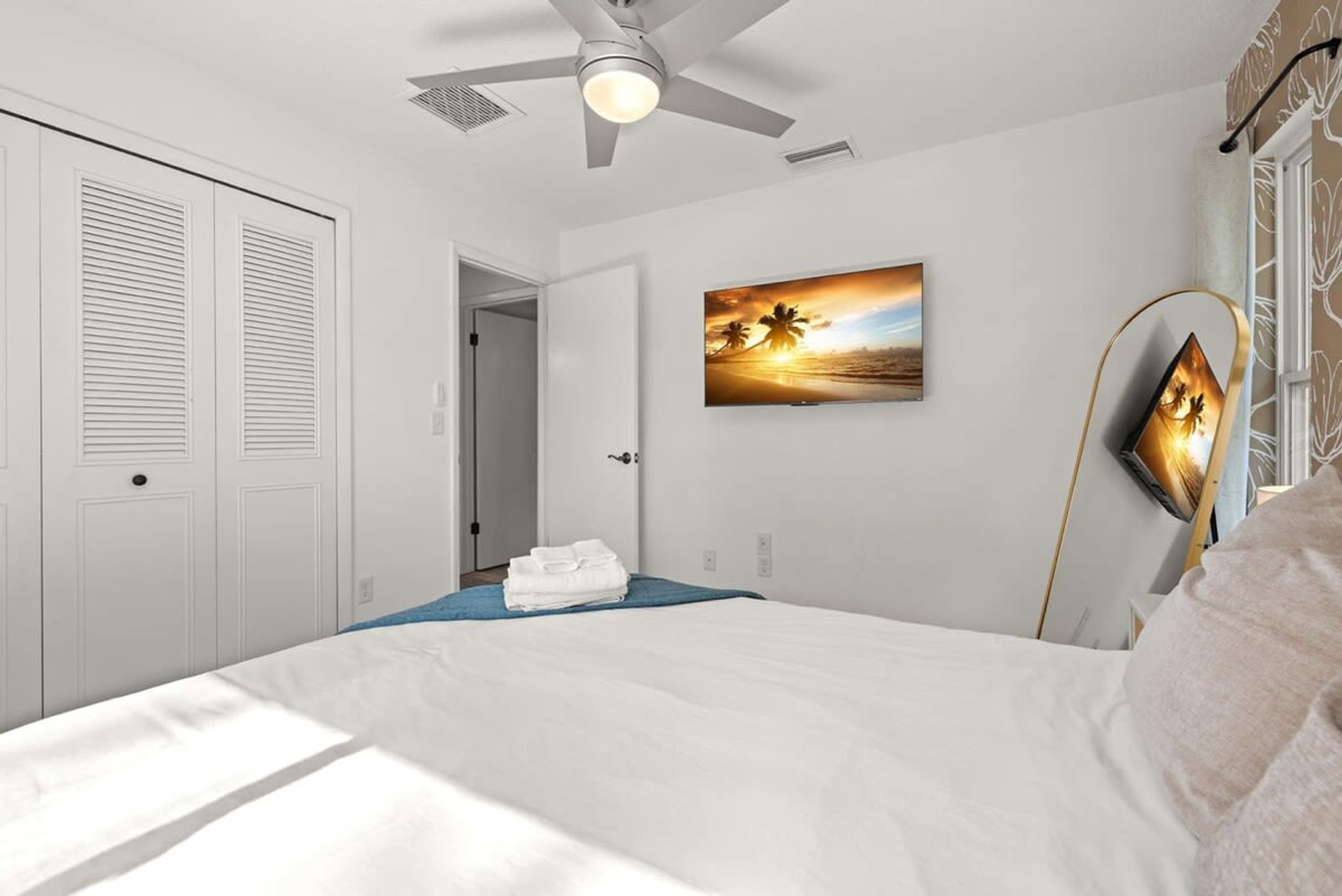 Um quarto com uma cama, um ventilador de teto, um quadro de um pôr do sol e uma porta.