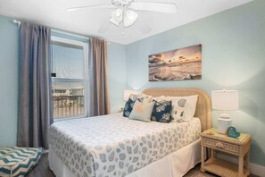 A bedroom with a bed, a nightstand, a lamp, a window with curtains, and a wall painting. - Ocean Views Boardwalk Access Large Community Pool (Port Aransas)