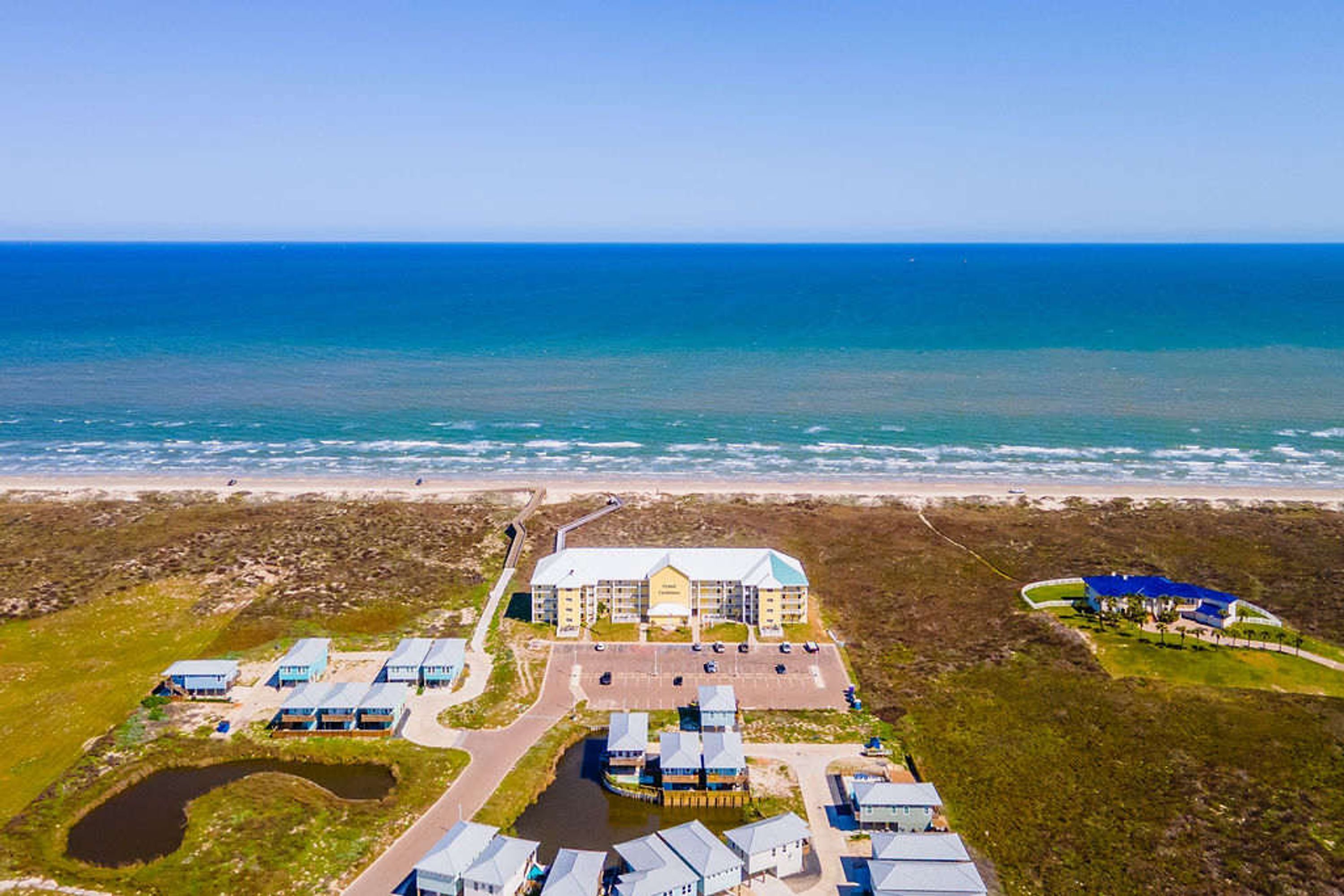 An aerial view of a coastal hotel with multiple buildings, a parking area, and a beach in the background.