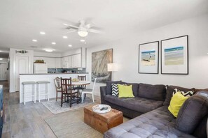 A modern living room with a gray sofa, a wooden coffee table, and a dining area with a white table and chairs. - Ocean Views Boardwalk Access Large Community Pool (Port Aransas)