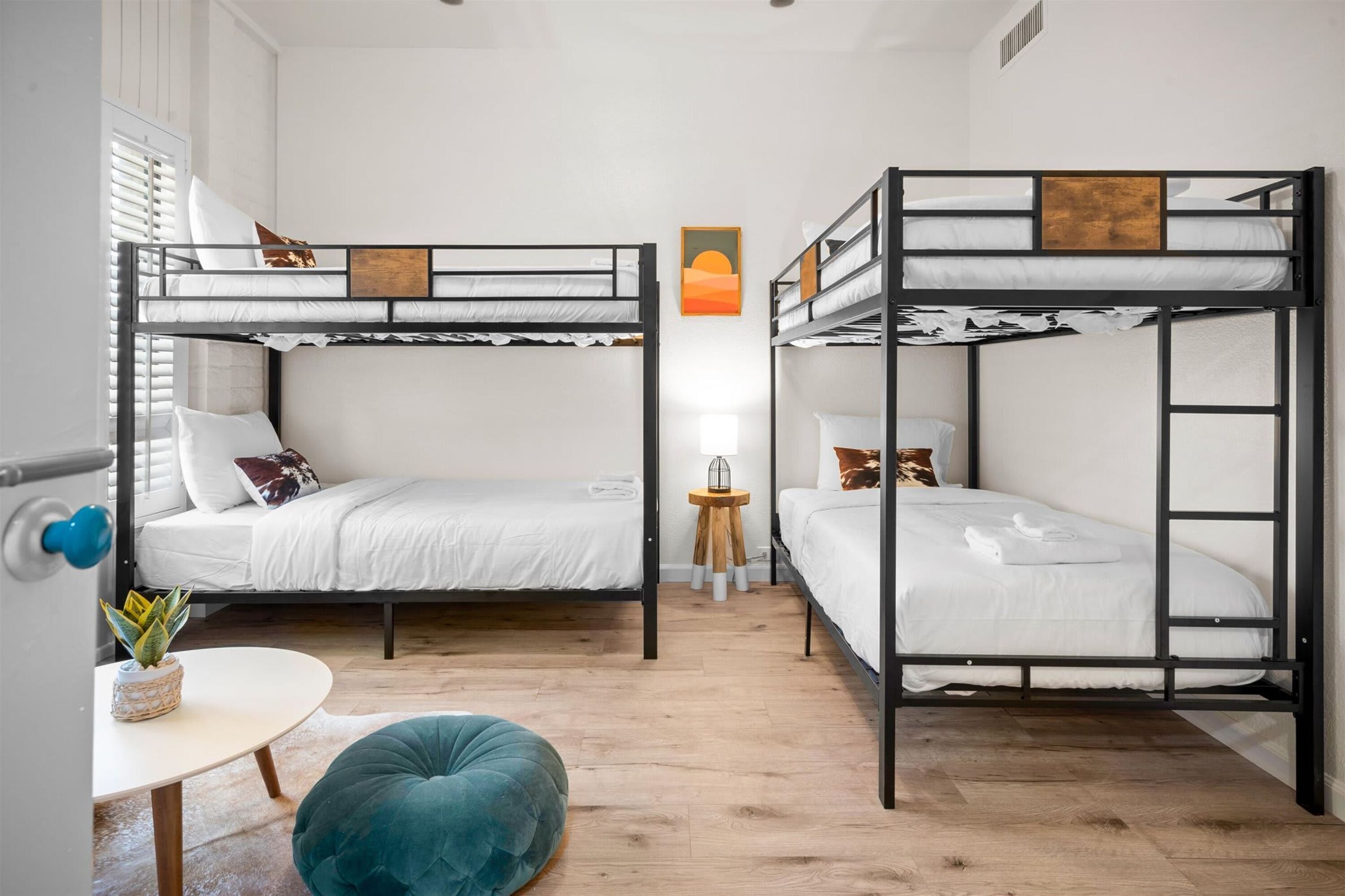 A room with two bunk beds, a small round table, a teal pouf, and a wall-mounted lamp.
