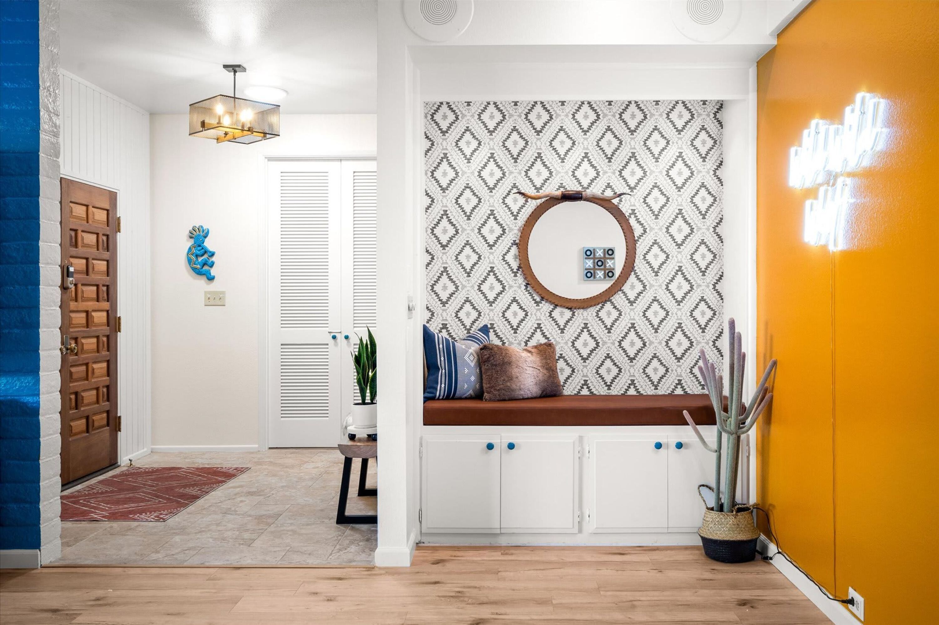 A hallway with a wooden door, a blue wall, a patterned wall, a round mirror, a bench with cushions, and a cactus plant.