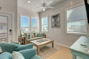 Ein Wohnzimmer mit einer Sofagarnitur, einem Couchtisch, einem Deckenventilator und großen Fenstern mit Jalousien.