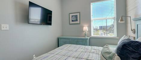A bedroom with a bed, a TV mounted on the wall, a window with blinds, a bedside table with a lamp, and a picture frame.